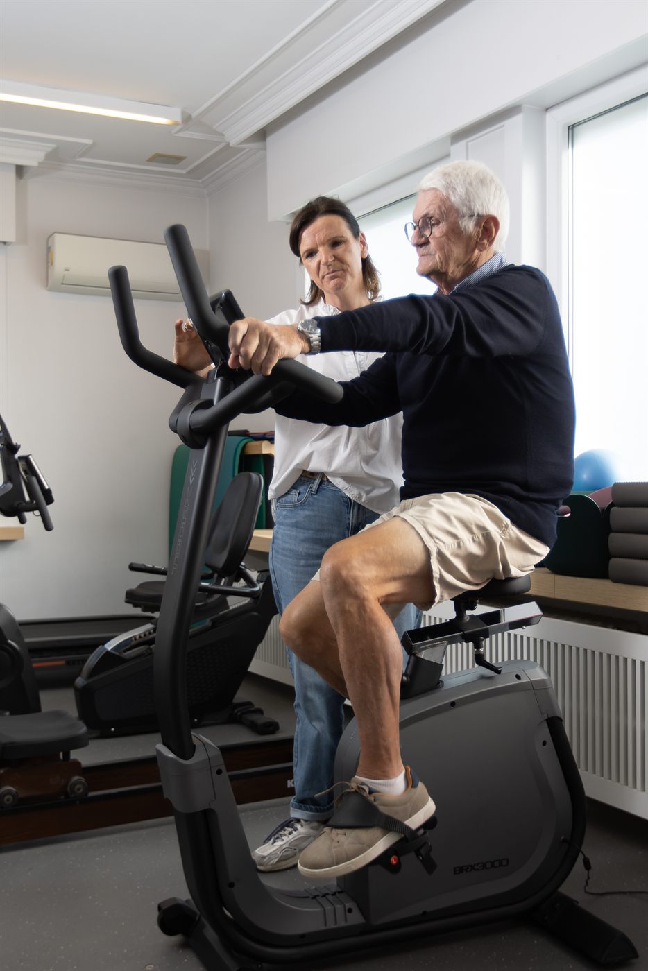 Oudere patiënt traint op de hometrainer onder begeleiding van de kinesitherapeute